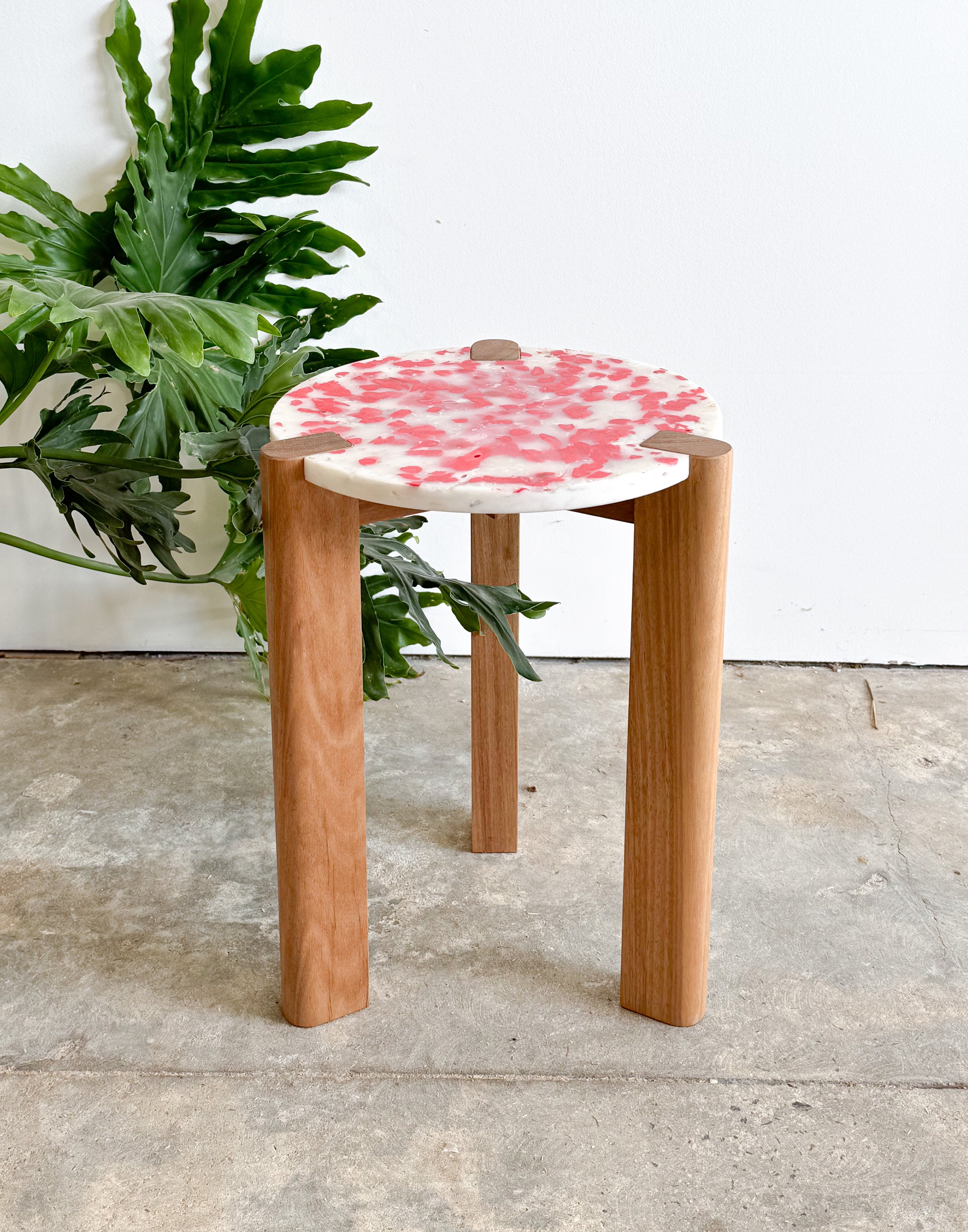 Short stool with 3 Tasmanian Oak timber legs and a red and white seat made from recycled soy sauce fish and milk bottles next to monstera